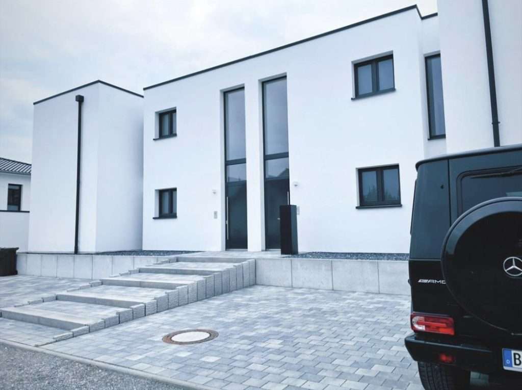 Weißes Mehrfamilienhaus mit Flachdach, hoher verglaster Hauseingang über einer Granittreppe, davor ein gepflasterter Vorplatz