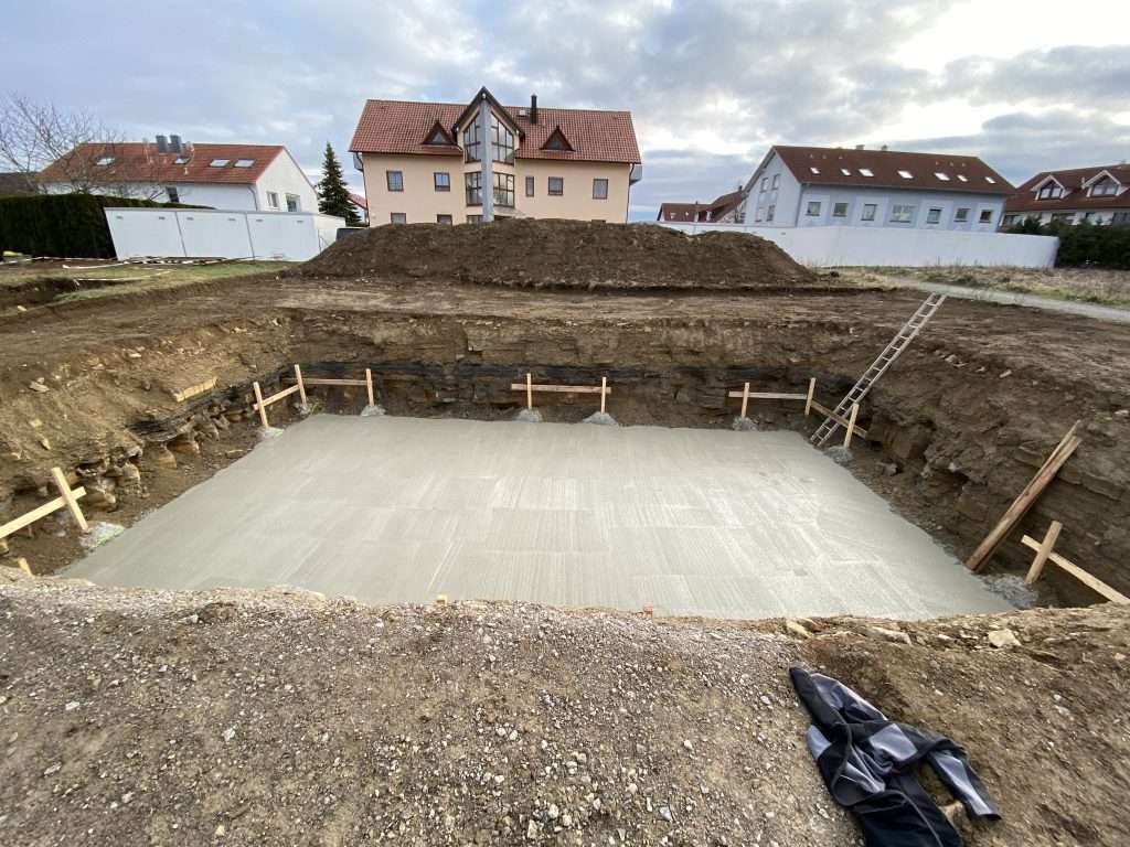 Ausgehobene Baugrube mit fertig betonierter Sohle und Schnurgerüst, dahinter die Wohnhäuser der Nachbarschaft
