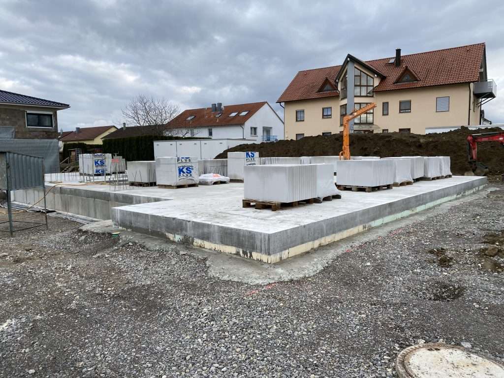 Fertige Bodenplatte des Neubaus, darauf Paletten mit Kalksandsteinen, dahinter ein Minikran und bestehende Wohnhäuser