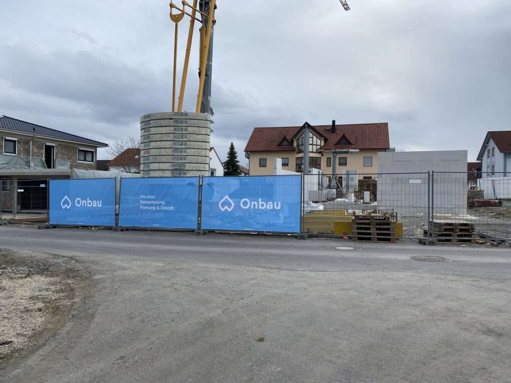 Baustelle hinter blauem Onbau-Bauzaun mit dem Schriftzug Neubau, Renovierung, Planung und Design, dahinter gestapelte Betonschachtringe und der Ausleger einer Betonpumpe