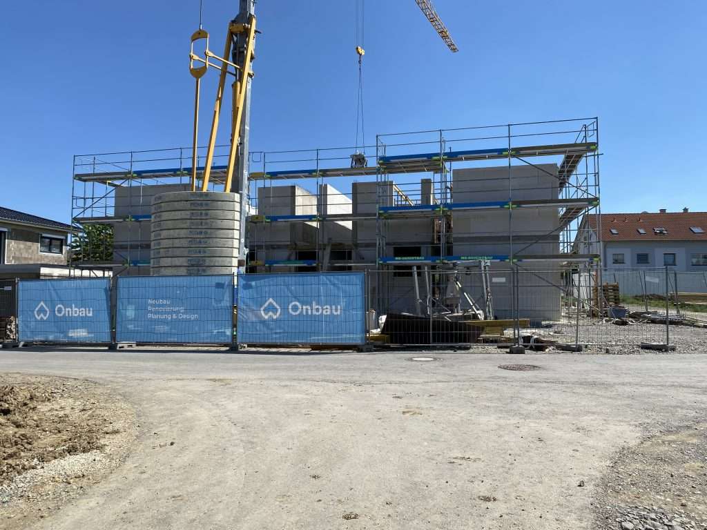 Rohbau des Mehrfamilienhauses mit Gerüst und Betonfertigteilwänden hinter dem Onbau-Bauzaun, daneben ein Baukran