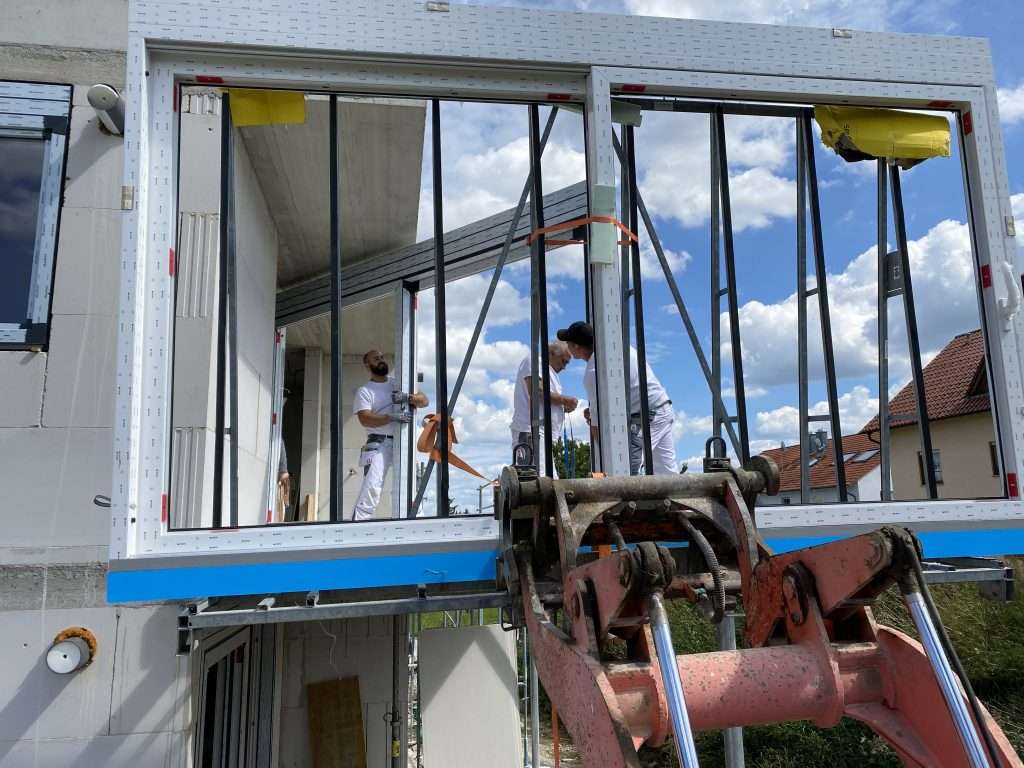 Drei Handwerker setzen ein großes Hebeschiebetür-Element in die Fassadenöffnung ein, gehalten von einem Teleskoplader