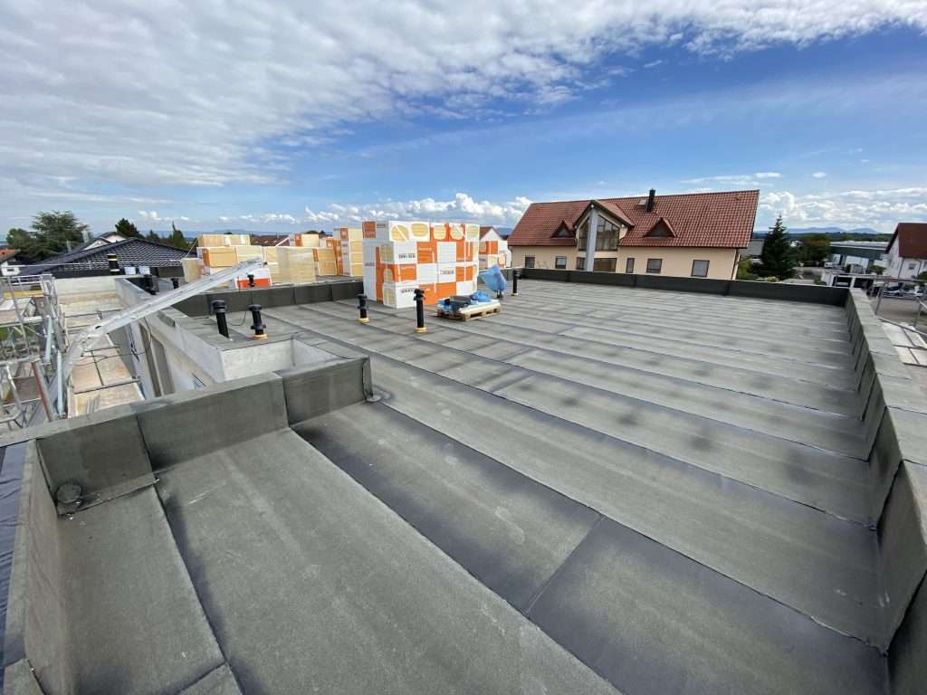 Flachdach des Neubaus mit verlegter Bitumen-Abdichtung, Attika und Dachabläufen, im Hintergrund gestapelte Dämmplatten